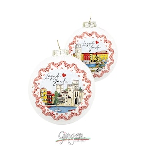 Può includere: Due palline di Natale bianche con illustrazioni del Lago di Garda, Italia. Ogni ornamento presenta un bordo decorativo rosso e la scritta "Lago di Garda". Le illustrazioni raffigurano edifici e monumenti colorati.