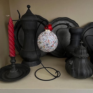 May include: A white and black patterned glass ornament with a red ribbon hanging from a black metal stand. The ornament is surrounded by black pewter serving dishes and a black candle holder with a red candle.