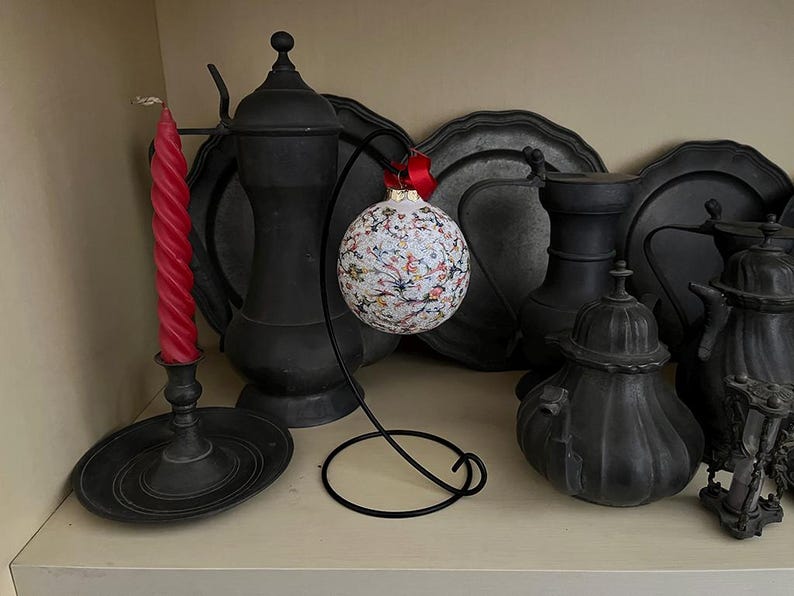 May include: A white and black patterned glass ornament with a red ribbon hanging from a black metal stand. The ornament is surrounded by black pewter serving dishes and a black candle holder with a red candle.