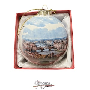 May include: A glass Christmas ornament with a painted image of the Ponte Vecchio bridge in Florence, Italy. The ornament is in a red box with white satin lining.