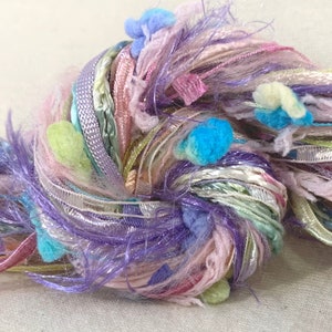 May include: A skein of colorful yarn with a mix of textures, including fluffy, silky, and ribbon-like strands. The yarn is a blend of pink, purple, green, and blue with small pom-poms throughout.