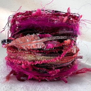 May include: A close-up of a bundle of pink, red, and gold yarn scraps. The yarn is arranged in a circular shape and has a fluffy, textured appearance.