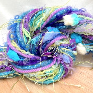 Puede incluir: Un ovillo de hilo colorido con una mezcla de texturas, incluyendo hebras esponjosas, brillantes y finas. El hilo es en tonos de azul, morado, amarillo y blanco.