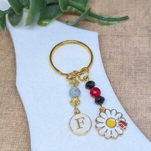 Peut inclure: Porte-clés en or avec un charme en forme de lettre F, un charme en forme de marguerite avec une coccinelle et une chaîne de perles colorées.