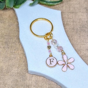 Könnte beinhalten: Goldener Schlüsselanhänger mit einem rosa Blumenanhänger und einem goldenen Anhänger mit dem Buchstaben F. Die Anhänger sind an einer goldenen Kette mit rosa und durchsichtigen Perlen befestigt.
