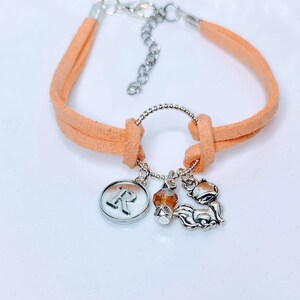 Op de afbeelding: Een perzik kleurige suède armband met zilverkleurige bedels. De bedels omvatten een ronde hanger met de letter "R", een oranje kristal en een kleine vos. De armband heeft een verstelbare sluiting.