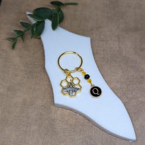 May include: Gold key chain with a bee charm in a honeycomb design, black and yellow beads, and a gold charm with the letter Q.