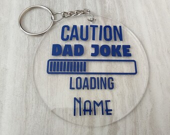 Op de afbeelding: Een transparante acryl sleutelhanger met een blauwe laadbalk en de tekst "CAUTION DAD JOKE LOADING NAME".