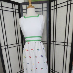 May include: A white sleeveless maxi dress with a green ribbon trim around the neckline and waist. The dress is embroidered with colourful flowers.