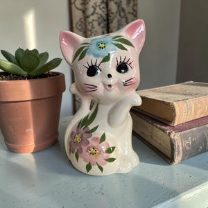 May include: A vintage ceramic cat figurine in a sitting pose. The cat is white with pink ears and floral accents in blue, purple, and pink. The cat has large black eyes and a smiling expression. A succulent plant in a terracotta pot is to the left.