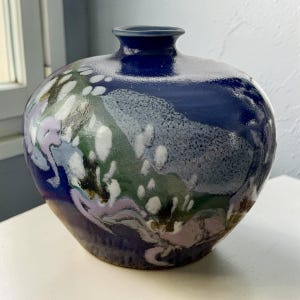 May include: A round ceramic vase with a dark blue exterior and a small opening at the top. The vase features a speckled design with white, green, and lavender accents. The vase has a glossy finish.