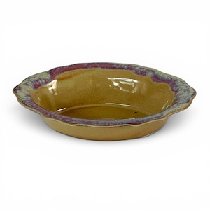 May include: A round ceramic pie dish with a smooth, tan-colored interior and a scalloped rim. The outer edge of the rim features a decorative glaze pattern with mottled shades of deep magenta, purple, and light blue, creating a textured appearance.