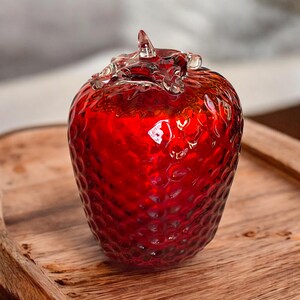 May include: A vibrant red glass strawberry with a textured surface and clear glass detailing on the top. The decorative piece is set on a wooden tray, showcasing its glossy finish and unique design. A charming accent for home decor.