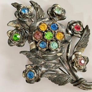 May include: A silver-toned metal brooch shaped like a bouquet of flowers. The piece features textured leaves and several individual rose-shaped blossoms, each centered with a faceted rhinestone in various colors including blue, green, pink, and yellow.