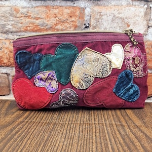 May include: A rectangular burgundy pouch featuring a zipper closure with a metallic pull. The exterior is decorated with various heart-shaped fabric appliqués in diverse textures, including solid red, dark green, blue, and patterned gold fabrics.