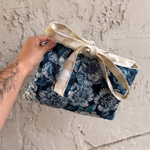 May include: A floral tote bag with a navy blue background and large blue and white flowers. The bag has a cream and tan gingham patterned trim and bow closure. The bag is held against a textured gray wall.