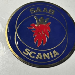 Könnte beinhalten: Ein rundes, blau-goldenes Emblem mit den Worten "SAAB" und "SCANIA" in goldener Schrift. Das Emblem zeigt in der Mitte einen roten Greif mit goldener Krone.
