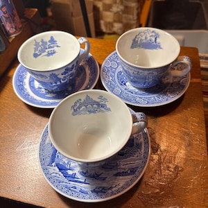 Peut inclure: Trois tasses et soucoupes bleues et blanches avec un motif de paysage. Les tasses et les soucoupes sont disposées sur une surface en bois. Les tasses ont des anses et les soucoupes ont une bordure détaillée.
