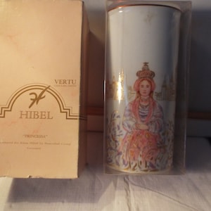 May include: A white porcelain vase with a gold trim and a painted image of a woman in a pink dress and crown. The vase is in a clear plastic box with the text "VERTU COLLECTION" and "HIBEL" on the box.