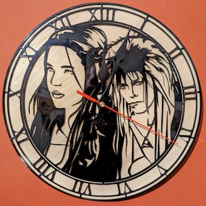 May include: A round wooden clock featuring a laser-cut design of three characters. The clock face has Roman numerals and black hands with a red second hand. The clock is made of light-colored wood with black accents.