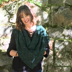 May include: A woman wearing a dark green shawl with a lace pattern, a black long-sleeved shirt, and blue jeans. She is standing in front of a stone wall with green moss.