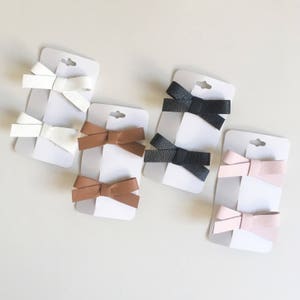 May include: A collection of hair bows in various colors, including white, brown, black, and pink. Each bow is made of a textured material and is attached to a small white card. The bows are arranged on a light-colored surface.