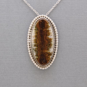 May include: An oval pendant necklace featuring a polished stone with brown, white, and green dendritic patterns. The stone is set in a silver bezel with a beaded edge, suspended from a delicate silver chain that attaches to the top of the pendant.