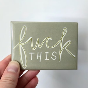 May include: A rectangular, sage green magnet with the words "fuck this" in a cursive, white font. The magnet is held by a hand against a white background. The text is in a playful, yet assertive style.