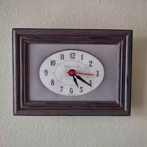 May include: A rectangular framed wall clock with an oval clock face. The clock face is white with black numbers and hands, and a red second hand. The frame is dark brown with a distressed finish. The clock is mounted on a textured, off-white wall.