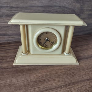 May include: A small, cream-colored mantel clock with a round clock face. The clock has a rectangular base and top, supported by two cylindrical columns. The clock face has black numerals and hands, with a gold-colored bezel.