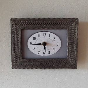 May include: An oval-faced clock with a silver-colored frame. The clock face is white with black numerals and hands. The frame has a detailed, textured design. The clock is mounted on a white wall.