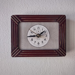 May include: A rectangular, vintage clock with a dark red frame and a white clock face. The clock face has black numerals and hands, with the words "WESTCLOX BABY BEN" printed at the top. The background is a light gray with a subtle pattern.