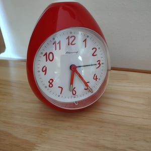 May include: A red egg-shaped alarm clock with a white face and black hands. The clock has a clear plastic cover and a white top. The clock face displays the word "Blessing". The clock is sitting on a wooden surface.