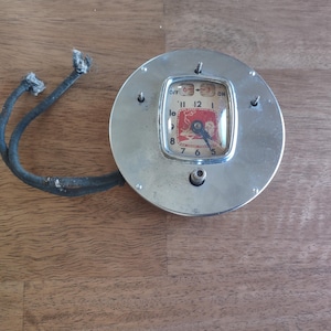 May include: A vintage clock with a silver-colored, circular casing. The clock face is rectangular with a red and white design. Two black cords extend from the side. The clock shows the time as 8:00.