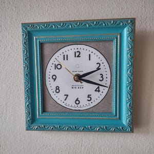 May include: A vintage wall clock with a white face, black numerals, and gold hands. The clock is framed in a turquoise, ornate rectangular frame with gold trim. The clock face reads "CHIME ALARM WESTCLOX BIG BEN".