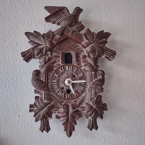 May include: A brown cuckoo clock with a bird perched on top. The clock features Roman numerals, two birds on the sides, and decorative leaves. The clock hands show the time.
