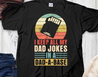 May include: Black t-shirt with the text "I KEEP ALL MY DAD JOKES IN A DAD-A-BASE". The design features a retro-style graphic of a data storage device set against a striped background in shades of teal, orange, and yellow.