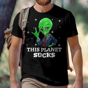 Puede incluir: Camiseta negra con un alienígena verde haciendo el gesto de la mano del rock and roll. El alienígena lleva una chaqueta azul y el texto "THIS PLANET SUCKS" está impreso debajo. La camiseta la lleva una persona con una mochila.