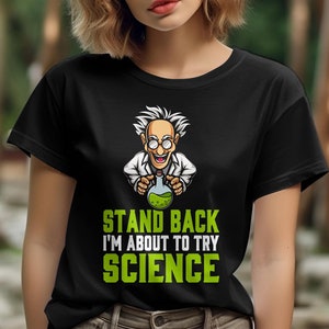 May include: Black t-shirt featuring a cartoon scientist holding a beaker with green liquid. The text "STAND BACK I'M ABOUT TO TRY SCIENCE" is printed in green and white. The shirt is made of a soft material.
