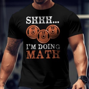 May include: Black t-shirt with a white graphic that says "SHHH... I'M DOING MATH" with three weight plates in orange with white numbers.