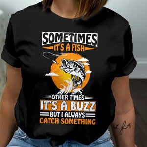 May include: Black t-shirt with a fishing-themed graphic. The design features a leaping fish, a fishing line, and a sunburst background. Text reads "Sometimes it's a fish, other times it's a buzz, but I always catch something."