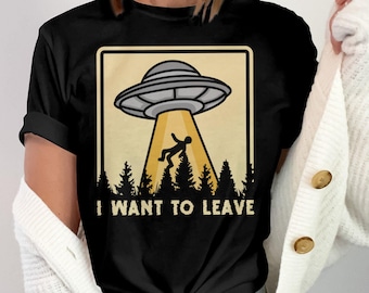 May include: Black t-shirt featuring a humorous graphic of a UFO beaming up a person above a forest silhouette. The text "I WANT TO LEAVE" is printed below the image in a bold, sans-serif font.