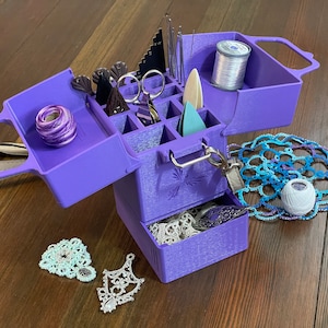 Puede incluir: Una caja de almacenamiento de plástico de color púrpura de varios niveles con un asa. La caja está abierta y llena de varios artículos de artesanía, incluidas agujas, hilo y un pequeño par de tijeras. También hay varios artículos pequeños de ganchillo blancos en la mesa.