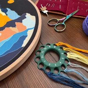 Könnte beinhalten: Ein Stickrahmen mit einem Landschaftsdesign mit Bergen und einem Mond. Ebenfalls sichtbar sind Stickschere, ein Fadenorganizer, ein Knäuel lila Garn und verschiedene Stickgarne in Blau-, Gelb- und Orangetönen. Ein laufendes Bastelprojekt.