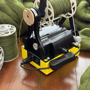 May include: A black and yellow yarn winder with two spools of green yarn. Two additional spools of green yarn are on the left. Green wool roving is in the background. The winder has a black power cord and a black on/off switch.