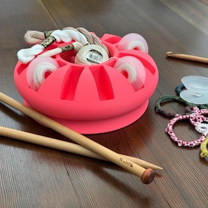 May include: A bright pink yarn organizer filled with white and beige thread spools and skeins. Two wooden knitting needles and a crochet hook are nearby, along with various crochet and knitting projects. A clear glass dish is also present.