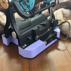 Può includere: Una filatrice elettrica nera e lilla con una ruota turchese e dettagli in legno. La macchina è collegata. Un gomitolo di filato colorato è dietro la macchina su una superficie di legno.