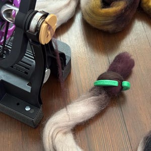 May include: A black and purple spinning wheel with a beige bobbin and a skein of fiber in shades of purple, brown, and beige. A green band secures the end of the fiber. The wheel sits on a wooden surface.