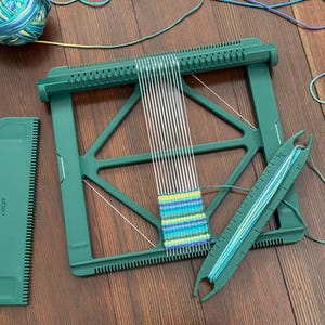 May include: A green weaving loom with white threads and a partially woven piece in blue and green. A green comb and a shuttle with blue yarn are also present. A ball of blue and green yarn sits nearby.