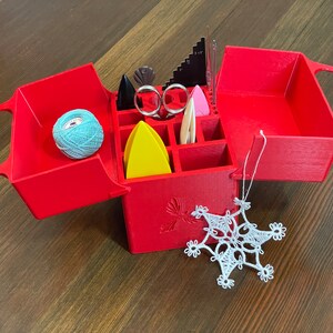 May include: Red plastic box with a hinged lid, divided into compartments. The box contains various craft supplies, including yarn, needles, and a small ruler. A white crocheted snowflake ornament is hanging from the box.